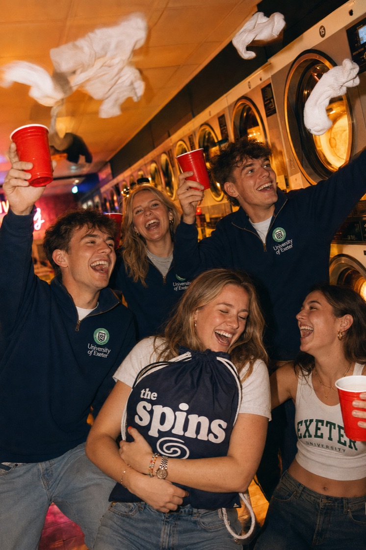 A group of students cheering with red cups raised at a laundromat party, holding a The Spins laundry bag