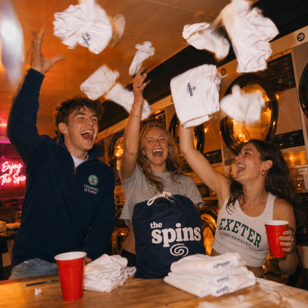 Students throwing clean clothes in the air like confetti at a laundromat party, red cups and a branded The Spins laundry bag