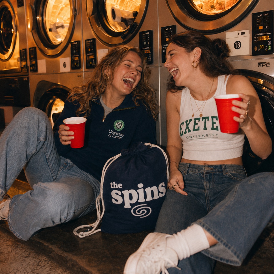 Two friends laughing with red cups in front of glowing washing machines and a The Spins laundry bag
