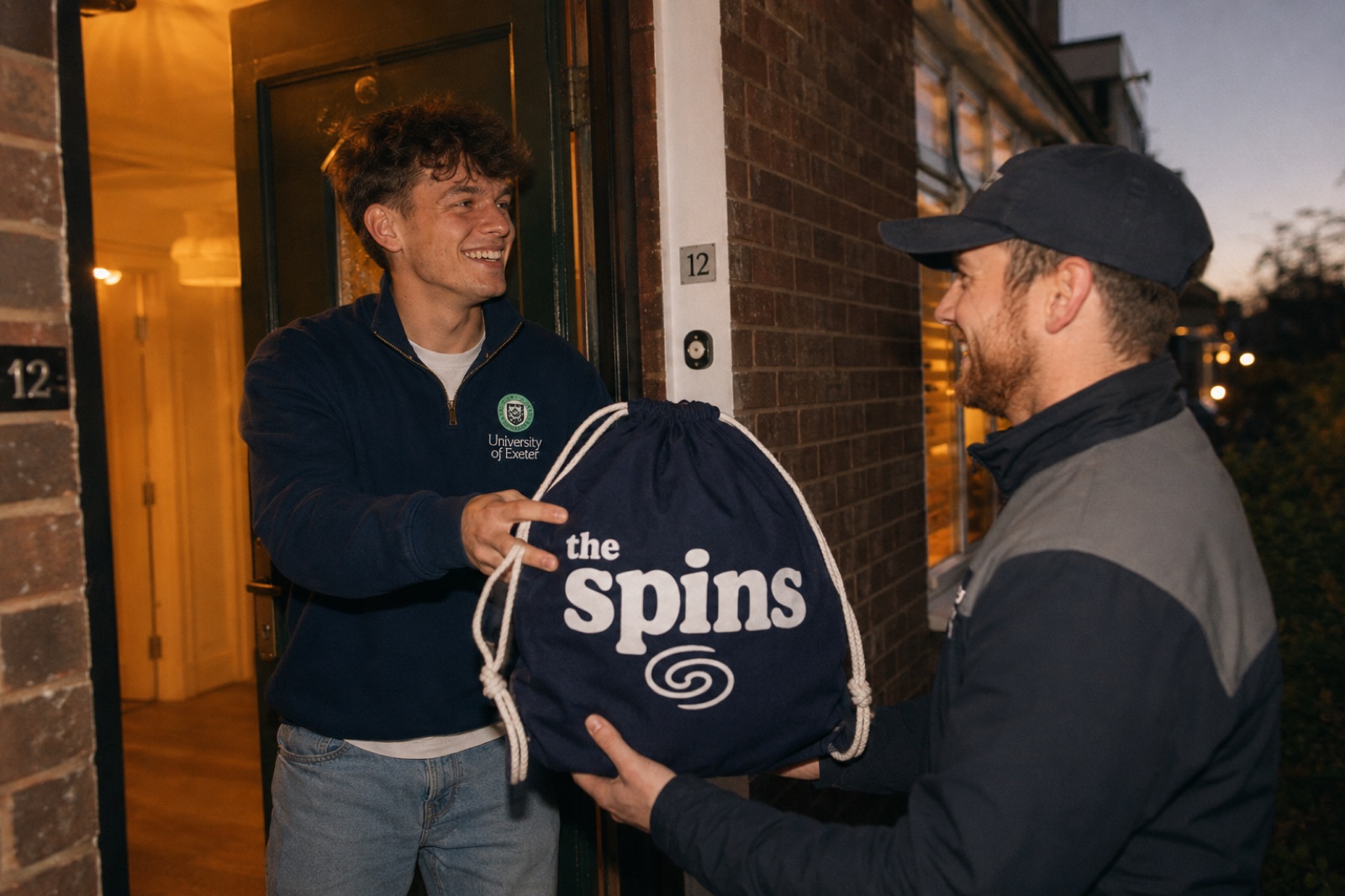 A student in a University of Exeter quarter-zip receiving a branded The Spins laundry bag from a delivery rep at their door at dusk